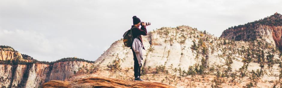 A photographer standing on a top of a rock taking pictures of mountain view