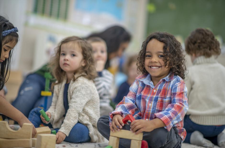 A super cute mixed race boy smiles at the camera as he is playing with some wooden blocks in his preschool classroom.