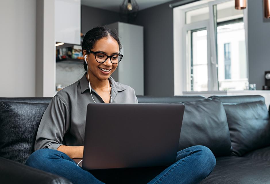 Young woman video calling using a laptop sitting on a sofa wearing earphones 